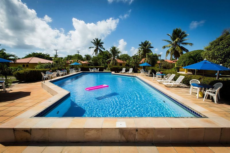 Piscina da pousada com espreguiçadeiras e guarda-sóis azuis, cercada de coqueiros e bangalôs amarelos